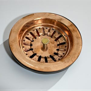 Puede incluir: Un colador de fregadero de color cobre con un diseño circular. El colador tiene una serie de pequeños agujeros espaciados uniformemente y un poste central con un pequeño pomo redondo de color latón. El borde es de color cobre más oscuro.