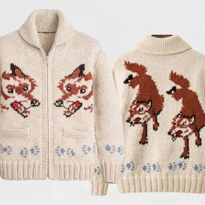 May include: Cream-coloured zip-up cardigan sweater with fox face and fox graphic designs. The sweater has a shawl collar, two front pockets, and blue floral accents on the cuffs and hem. The fox designs are brown, black, and red.