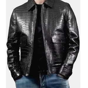 Könnte beinhalten: Schwarze Lederjacke mit Krokodilhautstruktur. Die Jacke hat einen Kragen, einen Reißverschluss und zwei Vordertaschen. Die Ärmel haben geknöpfte Manschetten. Die Person trägt eine blaue Jeans.
