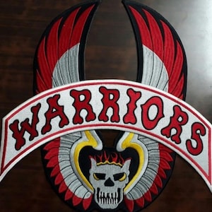 Puede incluir: Parche bordado con una calavera coronada, alas rojas, blancas, negras y amarillas. La palabra "WARRIORS" está arqueada en letras rojas sobre una pancarta blanca. El parche es para una chaqueta o chaleco.