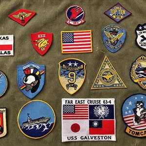 May include: A collection of military patches with various designs and text. Patches include an American flag, a Texas flag, and the text "DALLAS". Other patches feature eagles, ships, and the text "TOMCAT" and "USS GALVESTON".