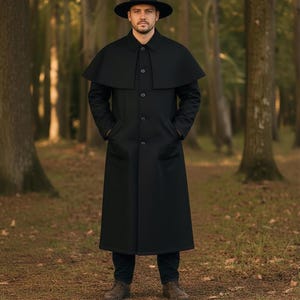 Puede incluir: Un abrigo negro largo con un diseño de hombros tipo capa y un sombrero negro a juego. El abrigo tiene cierre de botones y bolsillos. La persona está de pie en un bosque, rodeada de árboles y hojas caídas.