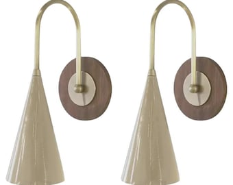 May include: Two wall sconces featuring a cone-shaped, cream-coloured shade and a curved brass arm. They are mounted on a round, dark wood backing with a brass centre.