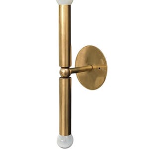 May include: A brass wall sconce with a minimalist design. It features two vertical cylindrical arms, each topped with a round, frosted glass bulb. The sconce is attached to a circular wall plate.