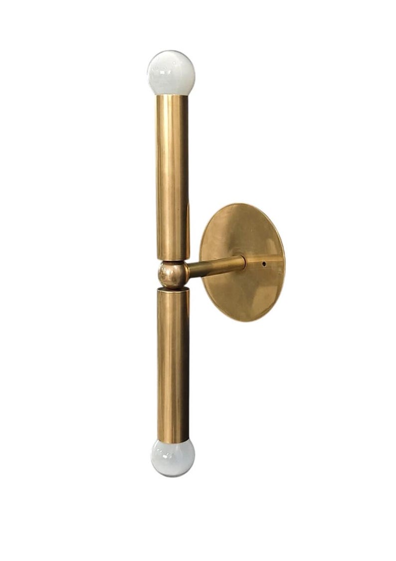 May include: A brass wall sconce with a minimalist design. It features two vertical cylindrical arms, each topped with a round, frosted glass bulb. The sconce is attached to a circular wall plate.