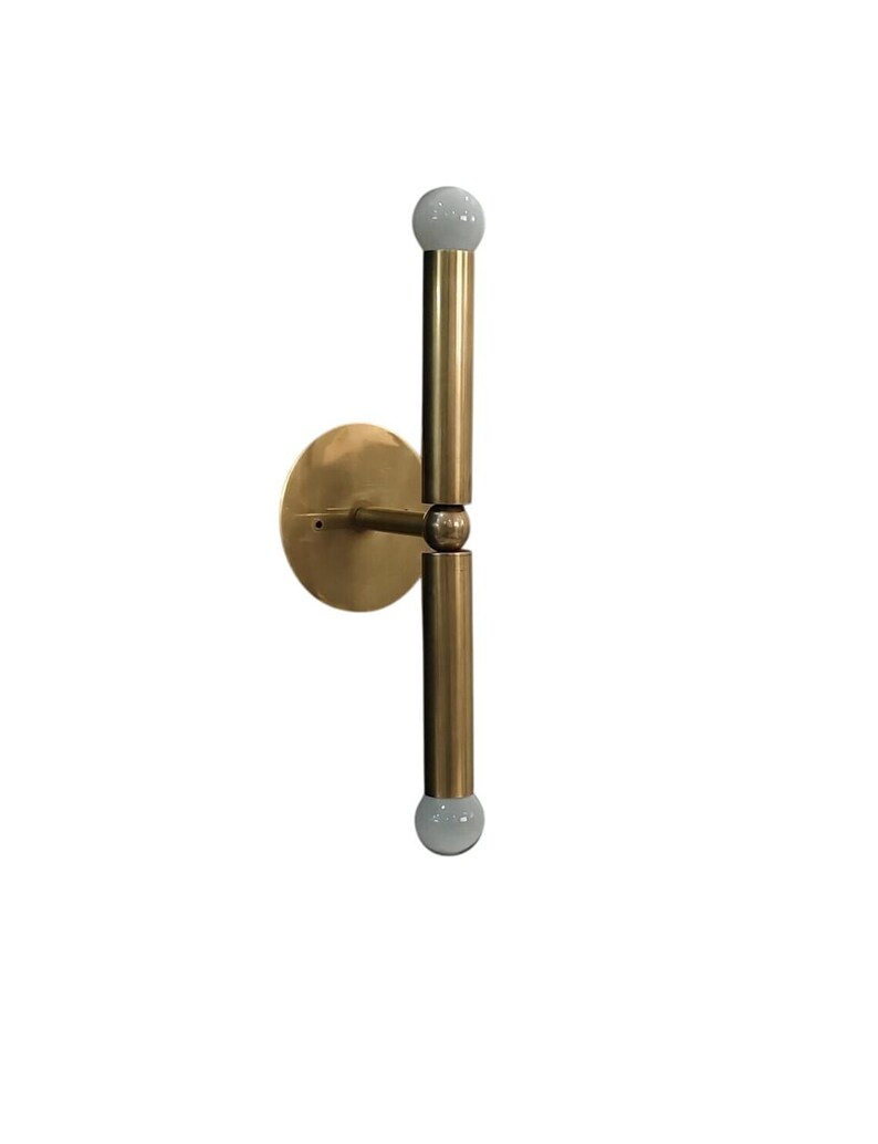 May include: A brass-colored wall sconce with a circular base and two vertical rods. Each rod has a white, spherical light at the end. The sconce is designed to be mounted on a wall and provides upward and downward lighting.