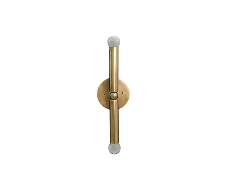 May include: A brass-toned wall sconce with a vertical design. It features a circular base with a central fixture and two parallel, cylindrical arms extending upwards and downwards. Each arm has a small, round, frosted glass bulb at the end.