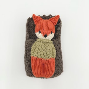 May include: A knitted fox doll wearing a green sweater and orange pants, lying in a brown knitted blanket.