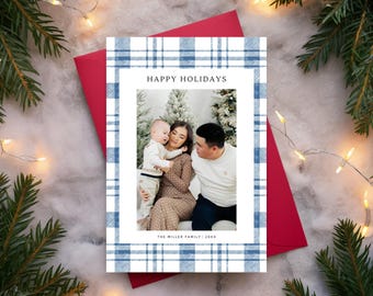 May include: A holiday card with a family photo inside a white frame, surrounded by a blue and white plaid pattern. The words "HAPPY HOLIDAYS" are at the top. The card is on a red envelope, with pine branches and fairy lights in the background.