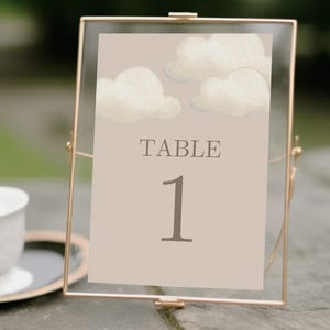 May include: A framed table number card with a gold frame. The card has a beige background with watercolor clouds and the word "TABLE" above the number "1". A white teacup and saucer are in the background.
