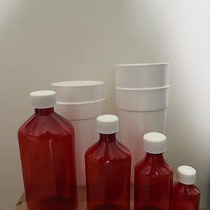 Könnte beinhalten: Eine Sammlung roter Plastikflaschen mit weißen Deckeln in verschiedenen Größen steht auf einer Kartonoberfläche. Im Hintergrund befinden sich zwei Stapel weißer zylindrischer Behälter. Die Flaschen sind leer und für Flüssigkeiten.