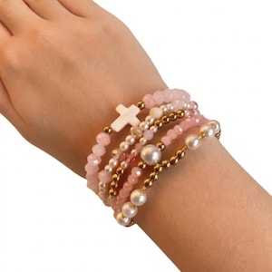 May include: A set of five stretch bracelets featuring a mix of light pink faceted beads, white faux pearls, and metallic gold-tone spherical beads. One bracelet includes a central white cross charm made of a smooth, opaque material.