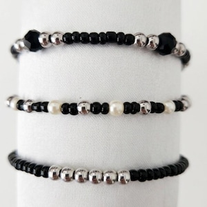 May include: Three beaded bracelets arranged vertically. The top bracelet features black faceted beads and silver-toned spheres. The middle bracelet alternates between black, silver-toned, and white pearl-like beads. The bottom bracelet combines black beads with a central row of silver-toned spheres.