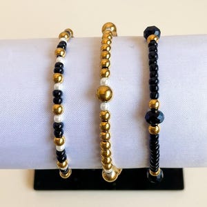 May include: Three beaded bracelets arranged side by side. The left bracelet features alternating black, white, and gold-toned beads. The centre bracelet consists primarily of gold-toned beads with small white accents. The right bracelet features black beads with gold-toned spacers.