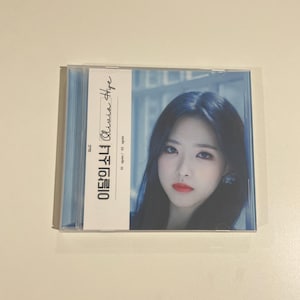 Puede incluir: Una caja de CD con una foto de una mujer con cabello oscuro y lápiz labial rojo. La caja tiene una etiqueta blanca con el nombre "Olivia Hye" en cursiva y texto coreano. El fondo es azul claro, en una funda de plástico transparente.