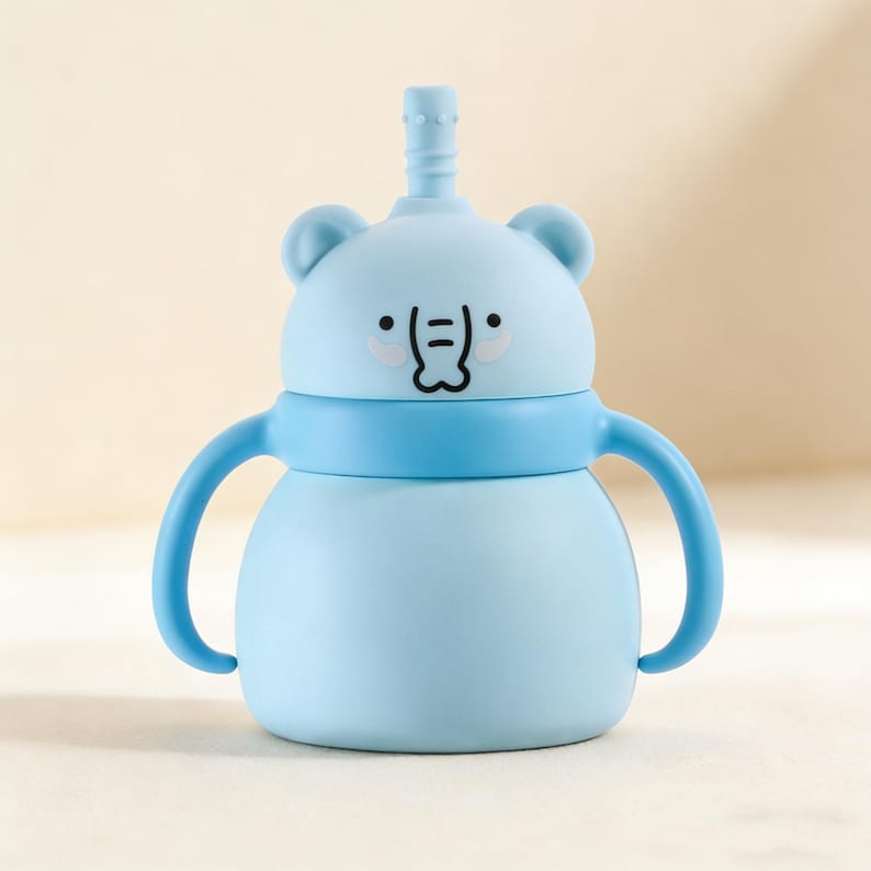 May include: A light blue baby sippy cup with a bear face design. The cup has a straw, two handles, and a blue band around the middle. The bear face has black eyes and a smiling mouth. The cup is designed for easy gripping.