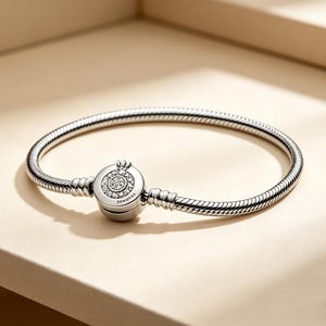 Op de afbeelding: Een zilveren armband met een ronde sluiting met een kroonontwerp en sprankelende heldere stenen. De armband heeft een slangenkettingontwerp en wordt gepresenteerd op een lichtbeige oppervlak.