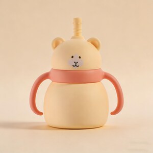 May include: A light yellow baby sippy cup with a bear face design. The cup has a pink band and handles, and a yellow straw. The bear face has a black mouth and two dots for eyes. The sippy cup is designed for babies and toddlers.