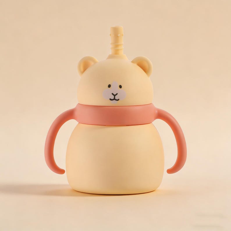 May include: A light yellow baby sippy cup with a bear face design. The cup has a pink band and handles, and a yellow straw. The bear face has a black mouth and two dots for eyes. The sippy cup is designed for babies and toddlers.
