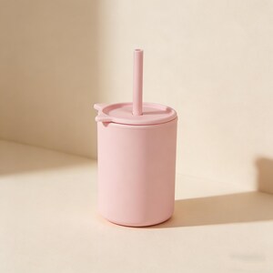 May include: A light pink sippy cup with a matching lid and straw. The cup is cylindrical with a rounded base. The lid has a small handle on each side. The straw is straight and extends upwards. The cup is made of a soft, matte material.
