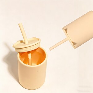 May include: A light peach-colored tumbler with a matching lid and straw. The lid is slightly ajar, revealing the interior of the cup. A second tumbler, also light peach-colored, is positioned nearby, with its straw extended. The items are made of a soft, matte material.