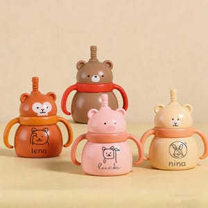 May include: Four colourful baby sippy cups with animal-themed designs. The cups feature bear, monkey, and pig shapes, with handles and a straw-like spout. The cups are in shades of brown, orange, pink, and yellow, with the names "lena", "luck", and "nina" printed on them.