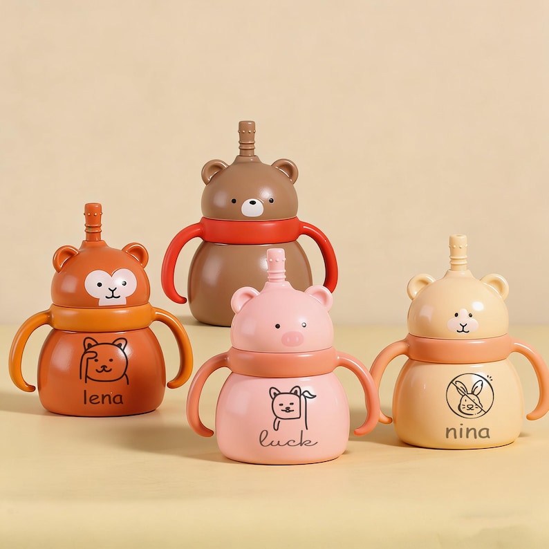 May include: Four colourful baby sippy cups with animal-themed designs. The cups feature bear, monkey, and pig shapes, with handles and a straw-like spout. The cups are in shades of brown, orange, pink, and yellow, with the names "lena", "luck", and "nina" printed on them.