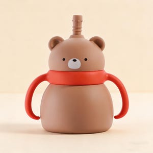 May include: A brown bear-shaped sippy cup with a red handle and a red band around the neck. The cup has a white nose and black eyes. The sippy cup has a straw and is designed for toddlers.