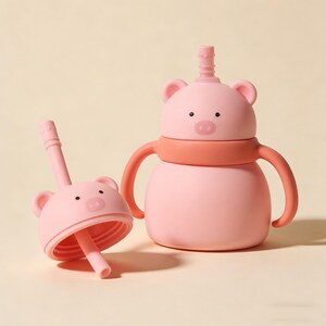 May include: A pink pig-shaped sippy cup with a matching lid and straw. The cup has a round body, two handles, and a pink band around the middle. The lid features a pig face with ears and a straw. The straw is removable.