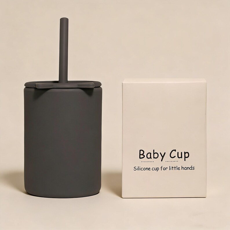 May include: A dark gray silicone baby cup with a straw and lid, next to a white box. The box has the words "Baby Cup" and "Silicone cup for little hands" printed on it.