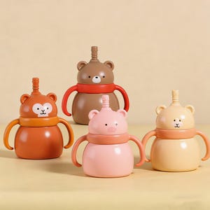 May include: Four colourful baby sippy cups with animal-themed designs. The cups feature a monkey, bear, pig, and bear design, each with a matching handle and straw. The cups are in shades of orange, brown, pink, and yellow.