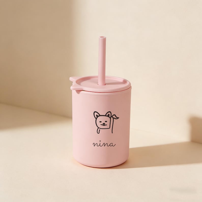 May include: A light pink tumbler with a matching lid and straw. The tumbler features a black illustration of a dog and the name "nina" in a script font. The straw is inserted into the lid.