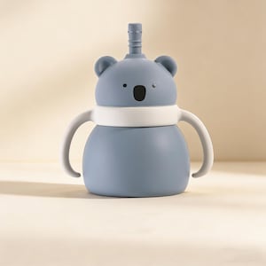 May include: A blue koala-shaped sippy cup with a white band and handles. The cup has a straw and a black oval-shaped mouth. The ears and eyes are also black. The cup is designed for toddlers and young children.