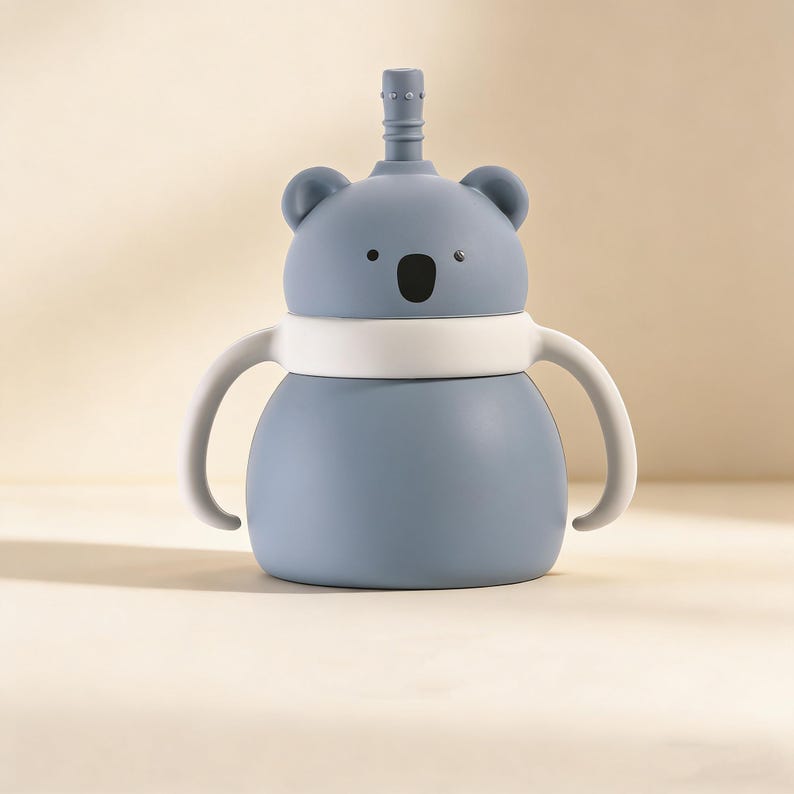 May include: A blue koala-shaped sippy cup with a white band and handles. The cup has a straw and a black oval-shaped mouth. The ears and eyes are also black. The cup is designed for toddlers and young children.