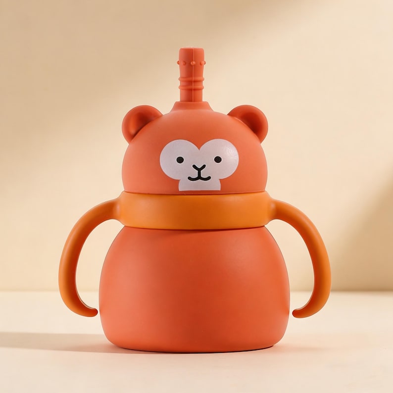 May include: Orange sippy cup with a monkey face design. The cup has a straw, two handles, and a white face with black eyes and a mouth. The cup is made of plastic and is designed for toddlers.
