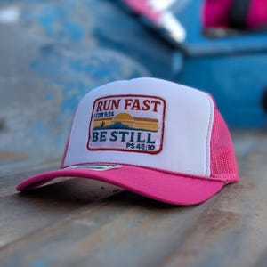Könnte beinhalten: Eine leuchtend pinke und weiße Trucker-Cap mit einem rechteckigen, gestickten Aufnäher auf der Vorderseite. Der Aufnäher zeigt den Text RUN FAST 1 COR 9:24 und BE STILL PS 46:10 sowie eine Grafik mit Sonne, Kaktus und Berglandschaft.