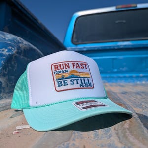 Op de afbeelding: Een witte en zeegroene truckerpet met de tekst "RUN FAST BE STILL" en bijbelverzen. De pet heeft een wit voorpaneel, een zeegroene klep en een mesh achterkant. De pet ligt op een blauw oppervlak.