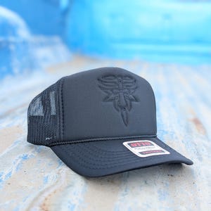 May include: Black trucker hat with a mesh back and a structured front panel. The front features an embroidered design. A small tag is attached to the brim. The hat is designed to be worn outdoors.