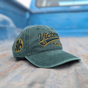 May include: A green baseball cap with the word "Victory" embroidered in gold script. The cap also features the numbers "33" and "316" in gold, with "IN CHARGE" embroidered on the side. The hat is made of a worn, textured fabric.