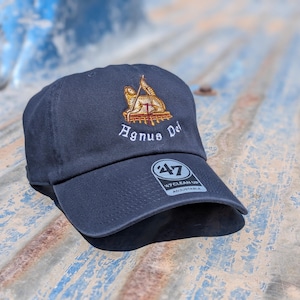 May include: A navy blue baseball cap featuring an embroidered gold and white Agnus Dei emblem above the text Agnus Dei. The cap includes a circular black and white 47 Clean Up adjustable sticker on the brim, showcasing a classic six-panel design.