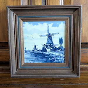 May include: A square wooden frame containing a blue and white ceramic tile depicting a landscape with two windmills, a small sailboat on the water, and a flock of birds in the sky. The frame has a tiered, stepped design with a dark brown finish.