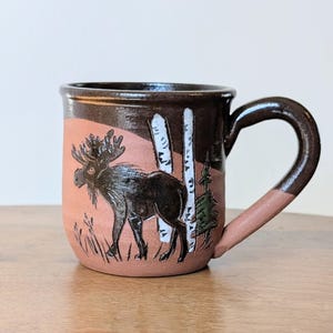 May include: A handmade ceramic mug with a brown handle and rim. The mug features a hand-painted design of a moose standing in a forest scene with birch trees and grass. The background is a light orange color.