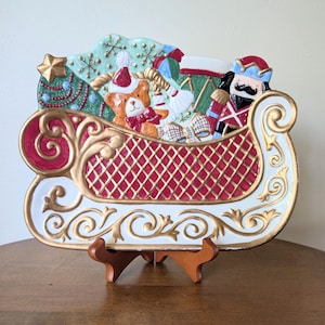 May include: A decorative Christmas sleigh-shaped plate with a red and gold lattice design. The plate is filled with festive items, including a teddy bear, a nutcracker, and a Christmas tree. The sleigh has a white and gold trim and is displayed on a wooden stand.