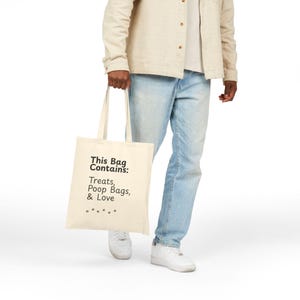 May include: Cream-colored tote bag with black text that reads "This Bag Contains: Treats, Poop Bags, & Love." The bag is held by a person wearing light-wash jeans and a tan jacket. The bag has a simple design and is made of a sturdy material.