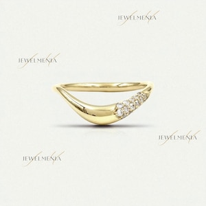 Puede incluir: Un anillo dorado curvado con un grupo de pequeñas piedras preciosas claras. El anillo tiene un acabado liso y pulido y un diseño moderno y minimalista. El fondo es de un color suave y neutro.