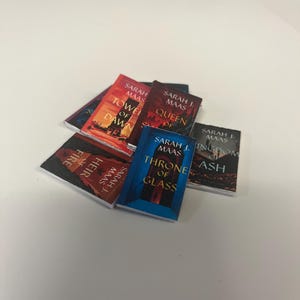 Può includere: Una collezione di libri in miniatura con copertine colorate. I titoli includono "Throne of Glass", "Queen of Shadows" e "Tower of Dawn" di Sarah J. Maas. I libri sono impilati su una superficie bianca.