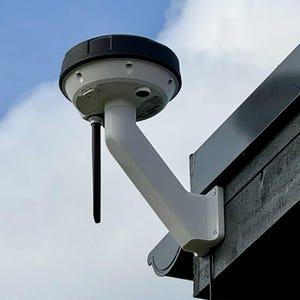 Op de afbeelding: Een wit en zwart buitenapparaat gemonteerd op een donkere houten constructie. Het apparaat heeft een cirkelvormige bovenkant en een gebogen arm, met een zwarte antenne. De achtergrond is een blauwe lucht met witte wolken.