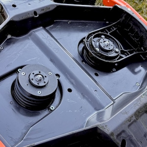 Op de afbeelding: Een close-up van de onderkant van een grasmaaierdek. Het zwarte dek heeft verschillende mechanische componenten, waaronder katrollen en riemen. Het dek heeft een oranje rand.