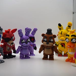 May include: A collection of colorful character figurines, including a gray robot, a red fox, purple rabbits, a brown bear, yellow figures, and a pink cupcake. The figures are standing on a white surface.