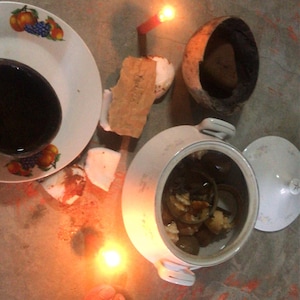 Puede incluir: Vista aérea de una escena con una vela encendida, un plato blanco con un líquido oscuro y una olla de cerámica con tapa. Se ve el pie de una persona con una sandalia. La escena está sobre una superficie de hormigón.